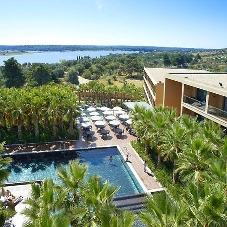 Ap Lago Montargil - Conference & Spa Buitenkant foto The photo shows a scenic view of a modern hotel or resort. In the foreground, there is a swimming pool surrounded by palm trees, with lounge chairs positioned beside it. Sun umbrellas provide shade over the chairs. The background features a lush land