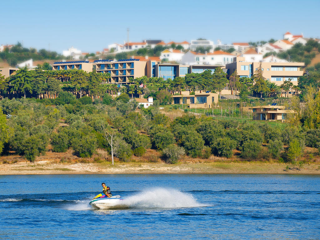 Ap Lago Montargil - Conference & Spa Buitenkant foto The photo depicts a person riding a jet ski on a body of water. In the background, there are modern buildings and lush greenery, suggesting a scenic landscape, likely near a resort or upscale residential area. The clear blue sky and calm water enhanc