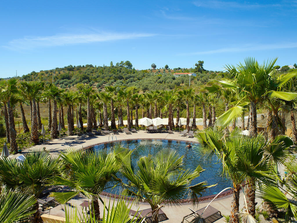 Ap Lago Montargil - Conference & Spa Buitenkant foto The photo depicts a beautiful outdoor area featuring a circular swimming pool surrounded by palm trees. The pool area has several lounge chairs arranged around it. In the background, there is a lush green hillside, and the sky is clear with a bright 