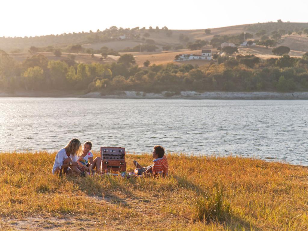 Ap Lago Montargil - Conference & Spa Buitenkant foto The photo depicts a serene lakeside scene during sunset. Three people are gathered on a grassy area near the water, enjoying a picnic. They are seated around a picnic basket, with the shimmering lake in the background and hills in the distance. The w