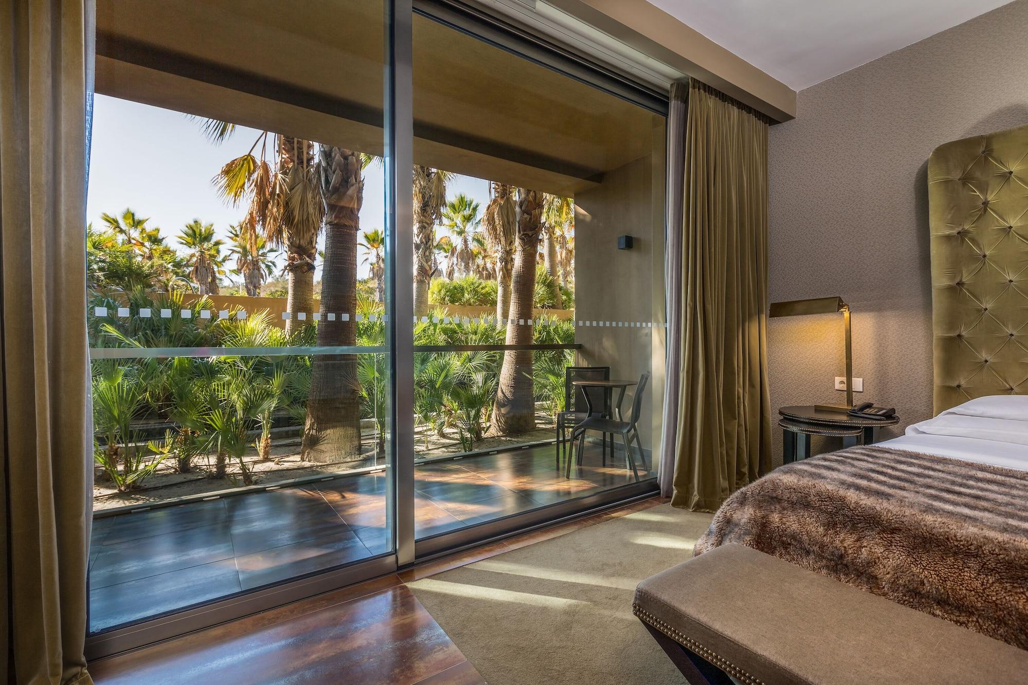 Ap Lago Montargil - Conference & Spa Buitenkant foto The photo shows a bedroom with large glass doors that lead out to a balcony or terrace. The view outside features palm trees and lush greenery, indicating a tropical or warm climate. Inside the room, there is a bed, a bedside table with a lamp, and a