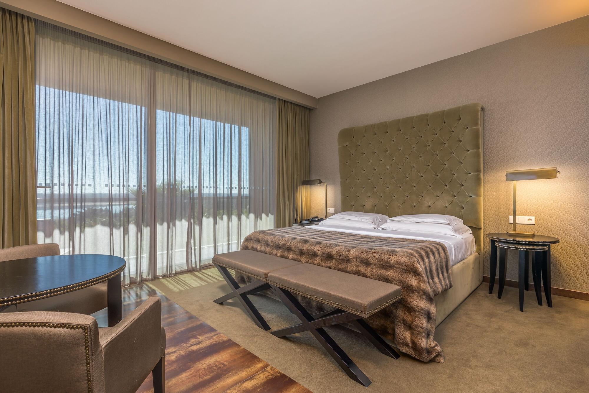 Ap Lago Montargil - Conference & Spa Buitenkant foto The photo shows a modern hotel room featuring a large bed with a tufted headboard and a cozy blanket. There are curtains draped over large windows that allow natural light to flow in, providing a view of the outside. A small seating area includes two