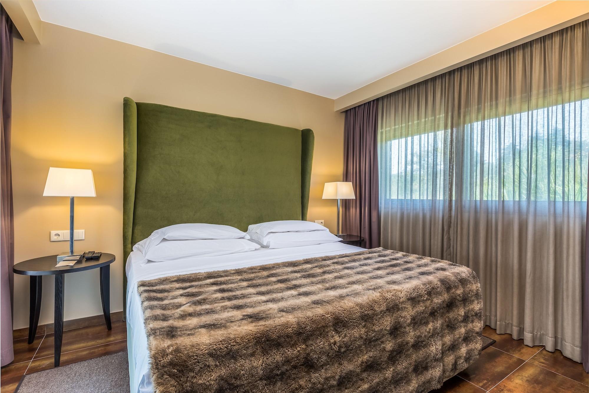 Ap Lago Montargil - Conference & Spa Buitenkant foto The photo shows a cozy hotel room featuring a large bed with two white pillows and a fur-style blanket draped over it. Behind the bed is a tall, green headboard that adds a touch of color to the room. On either side of the bed are bedside tables, eac