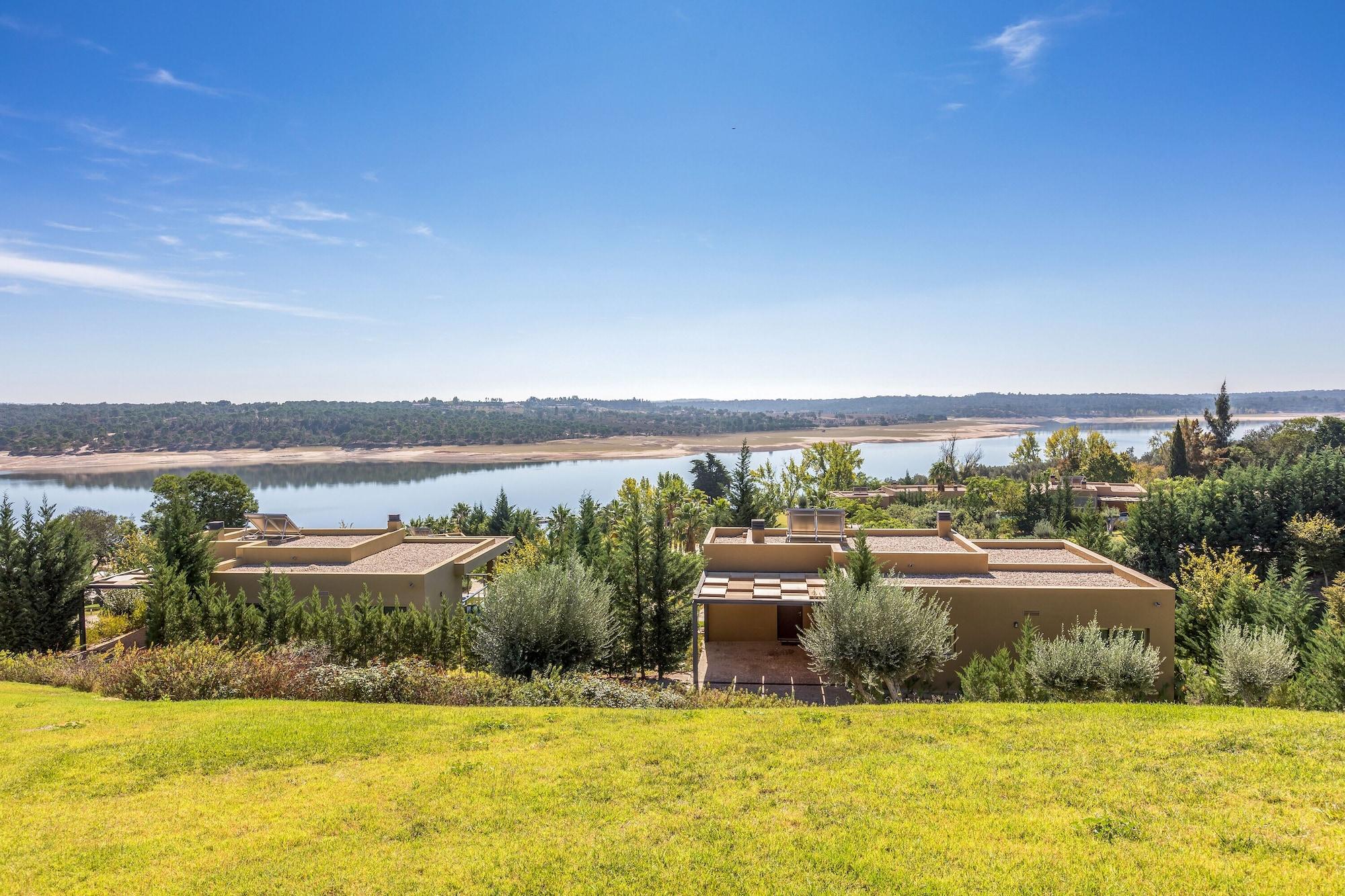 Ap Lago Montargil - Conference & Spa Buitenkant foto The photo depicts a scenic landscape featuring a calm body of water, possibly a lake or river, surrounded by lush greenery and trees. In the foreground, there are several modern buildings with flat roofs, set against a vibrant blue sky. The overall s