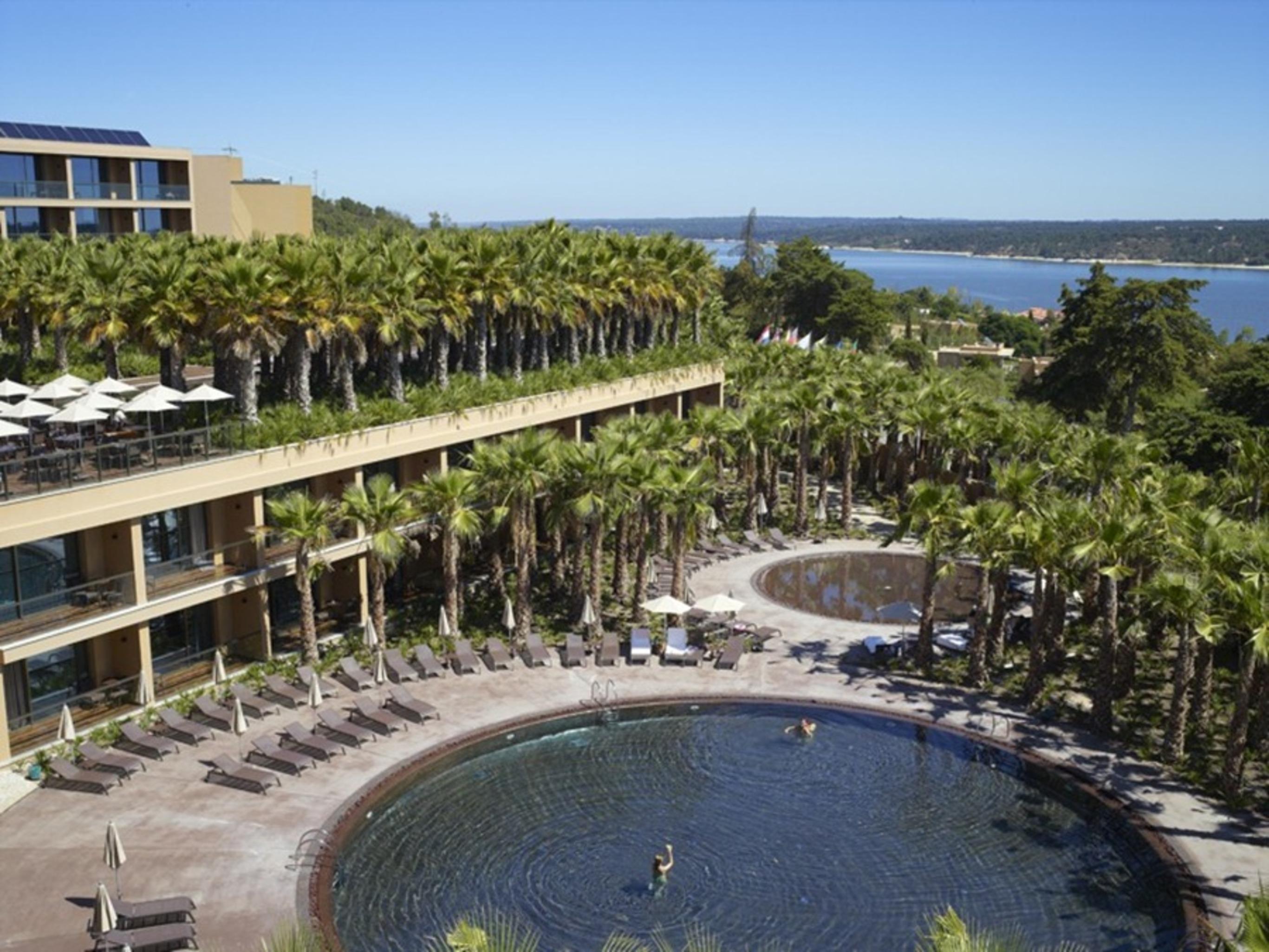 Ap Lago Montargil - Conference & Spa Buitenkant foto The photo shows a scenic view of a resort area. In the foreground, there are circular pools surrounded by lounge chairs, with a few people enjoying the water. Palm trees line the edges, creating a tropical atmosphere. In the background, there is a mu