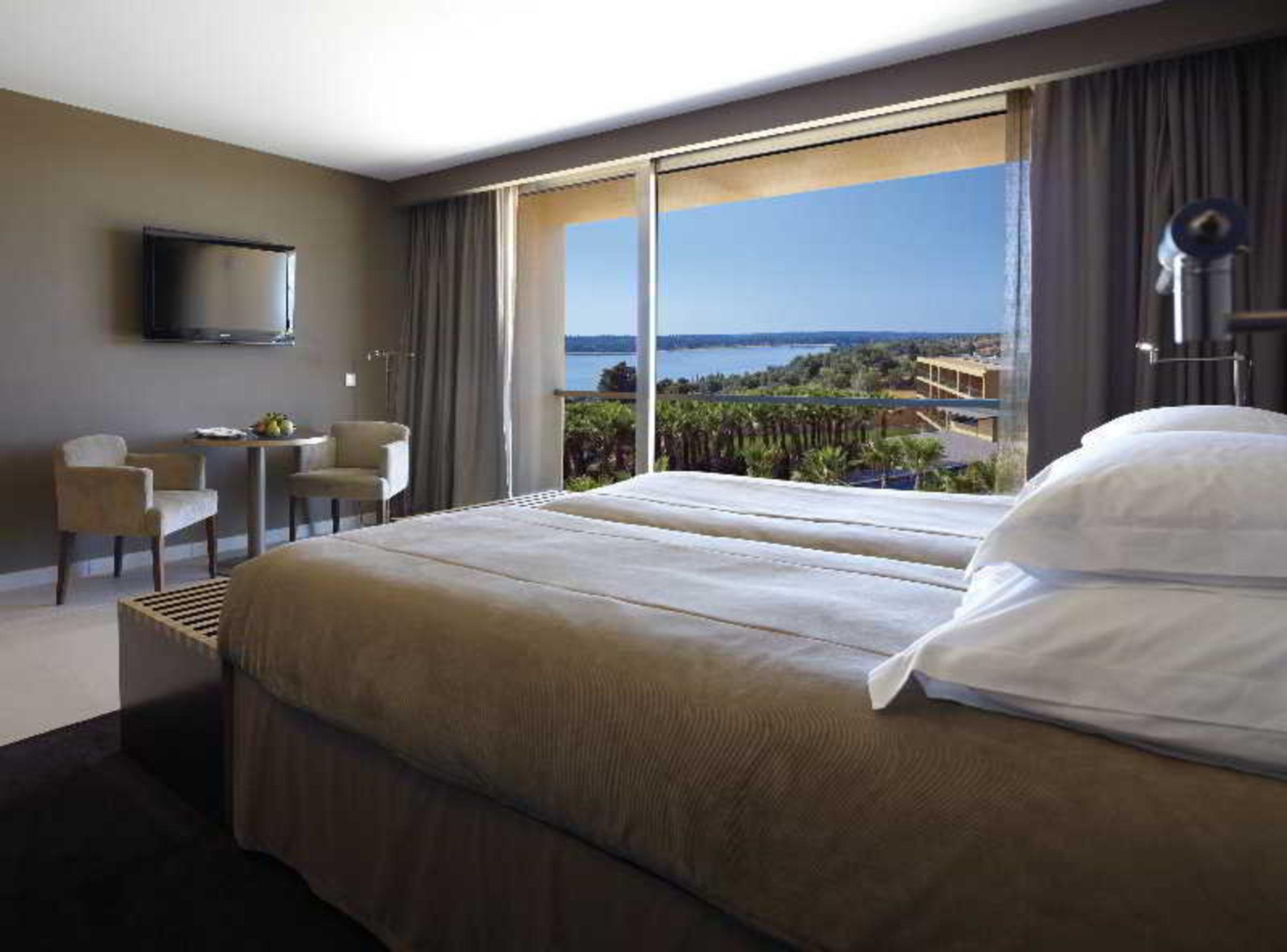 Ap Lago Montargil - Conference & Spa Buitenkant foto The photo shows a modern hotel room with a large window offering a view of the sea and mountains. The room features a comfortable bed with white pillows and brown bedding. There is a small sitting area with two chairs and a table, as well as a wall-m