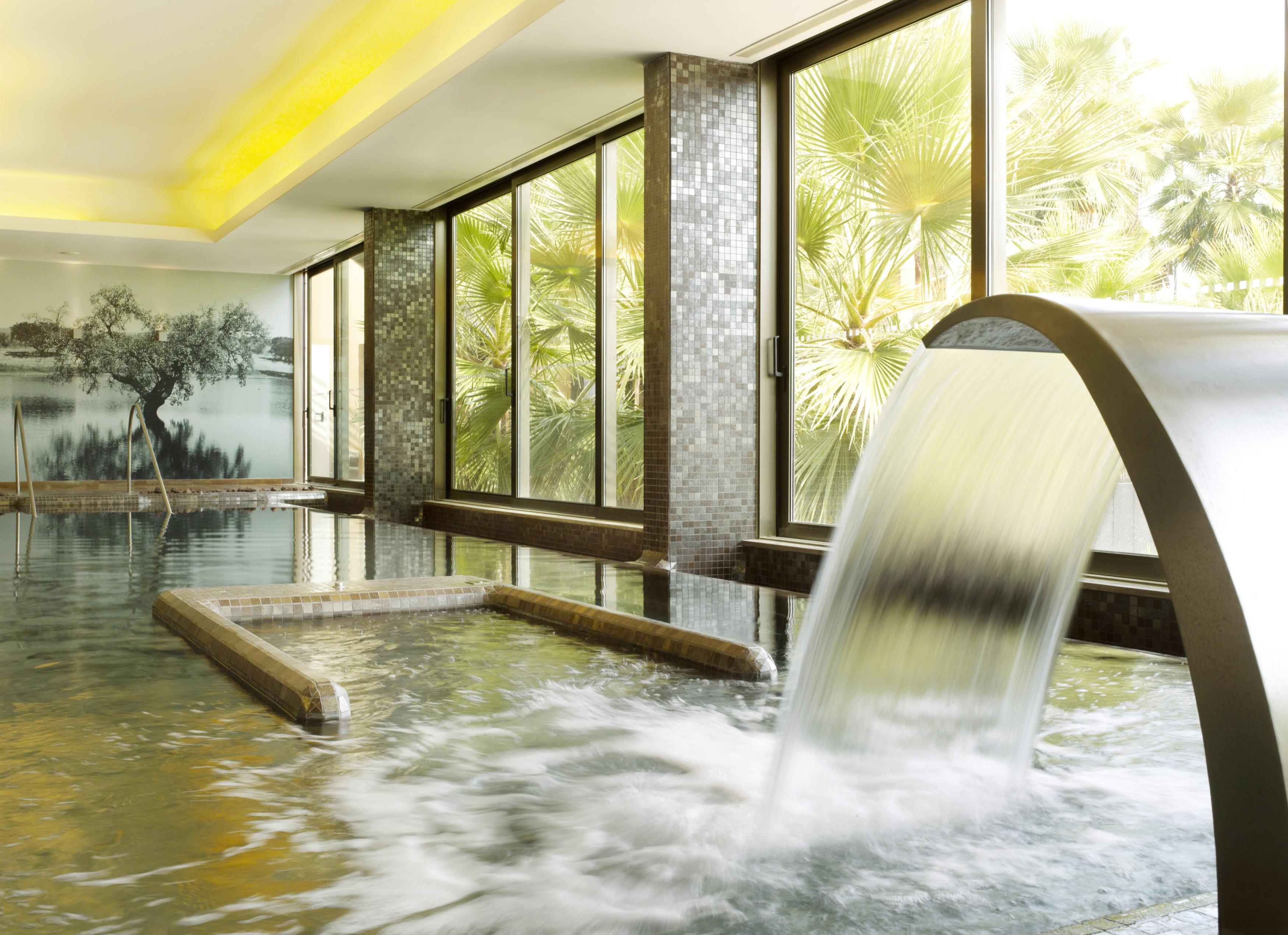 Ap Lago Montargil - Conference & Spa Buitenkant foto The photo depicts a serene spa environment featuring a tranquil indoor pool area. There is a sleek, modern water feature that spills water gracefully into the pool. Large windows allow natural light to flood the space, showcasing views of lush greene