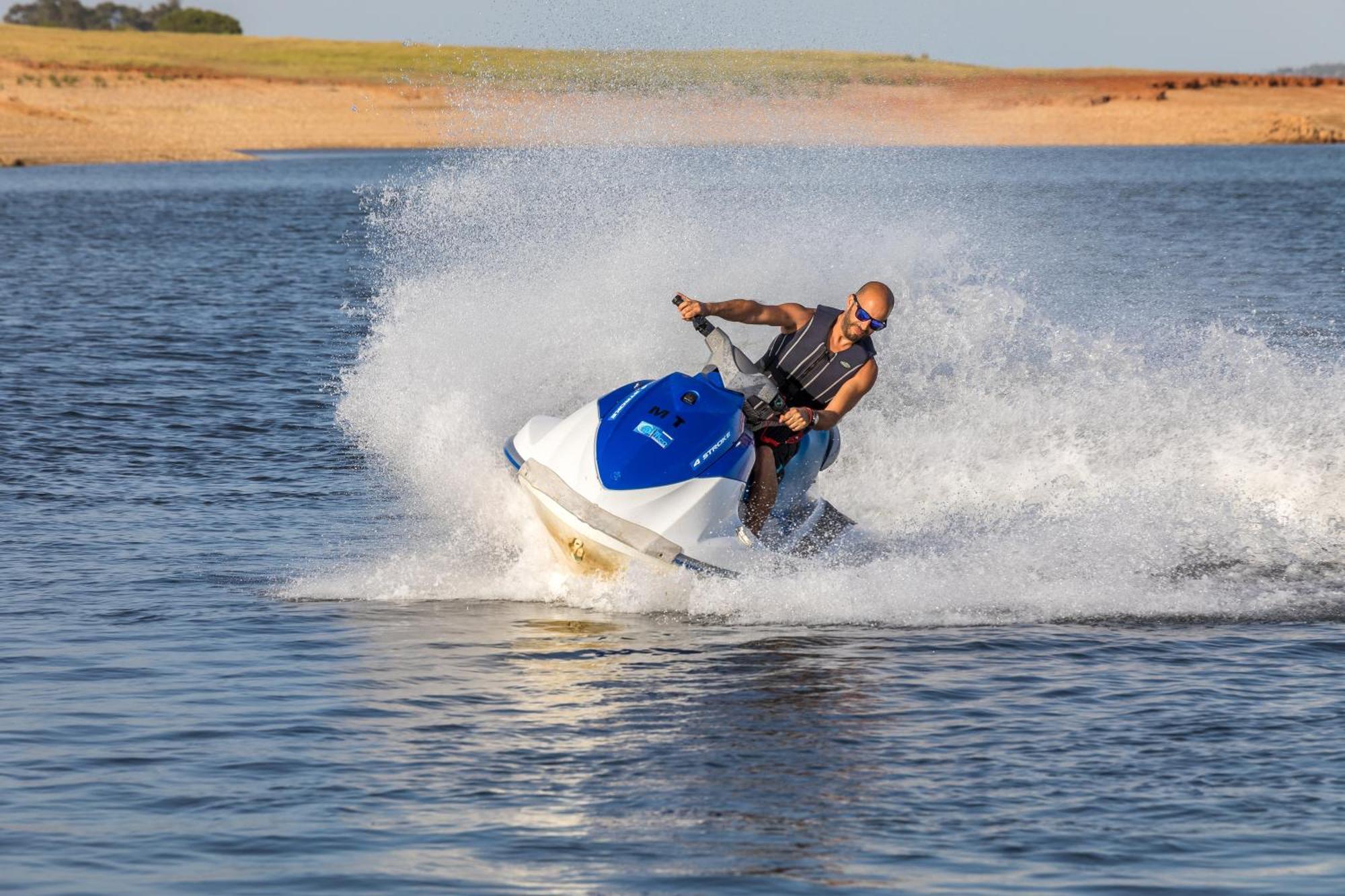 Ap Lago Montargil - Conference & Spa Buitenkant foto The photo shows a person riding a jet ski over a body of water. The rider is wearing sunglasses and a life vest, leaning forward as the jet ski accelerates, creating a spray of water around them. The background features a natural landscape, likely a 