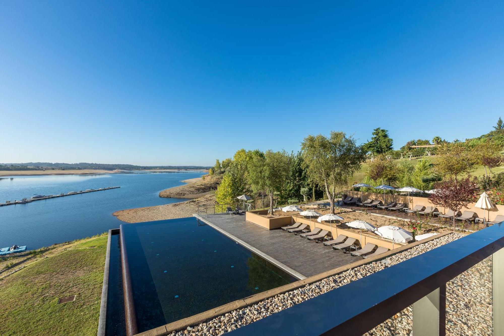 Ap Lago Montargil - Conference & Spa Buitenkant foto The photograph depicts a scenic view of a tranquil water body, likely a lake or river, bordered by lush greenery. In the foreground, there is an infinity pool that blends seamlessly with the water beyond. Comfortable lounge chairs are arranged on a w