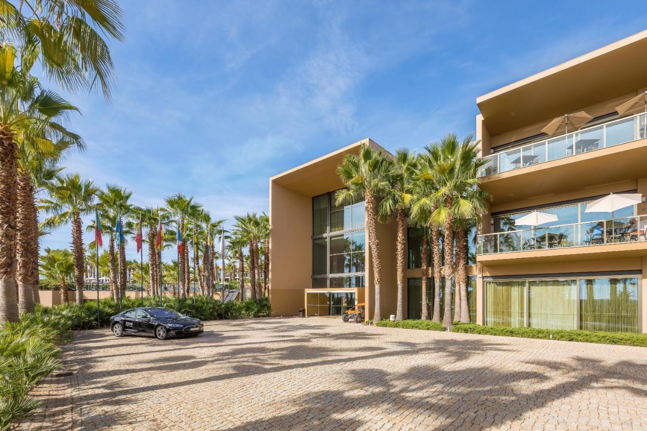 Ap Lago Montargil - Conference & Spa Buitenkant foto The photo shows a modern building with a sleek design, featuring large glass windows and balconies. The structure is surrounded by a well-maintained outdoor area with palm trees, adding a tropical feel. In front of the building, there is a paved park